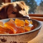 Close-up of golden Apple & Bone Broth Gummy Bones for Dogs in a ceramic bowl, with a dog looking on.