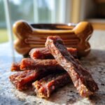 Close-up of delicious Chicken Pear Smooth Jerky sticks piled on a granite surface with a bone-shaped dish in the background.
