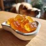 Amber-colored Ginger Bone Broth Tummy-Soothing Gummies in a small dish with a dog looking on in the background.