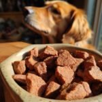 Close-up of Homemade Air Dried Beef and Sweet Potato Kibble Bites in a ceramic bowl with a dog looking eagerly in the background.