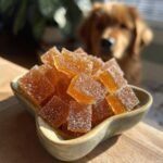 A bowl of amber-colored Soft Chew Bone Broth Training Gummies for Dogs with a golden retriever looking on in the background.
